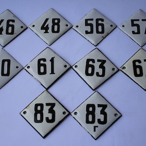 May include: Ten white diamond-shaped porcelain house number plaques with black numbers. The numbers are 46, 48, 56, 57, 60, 61, 63, 67, 83, and 83.