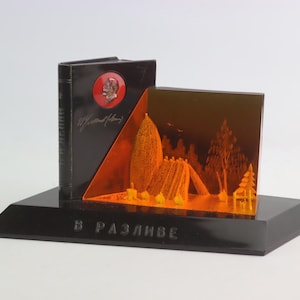 Può includere: Una scultura in vetro nero e arancione che rappresenta una scena di bosco con un libro che presenta un profilo rosso di un uomo sulla copertina. La base della scultura è nera e recita "В РАЗЛИВЕ".