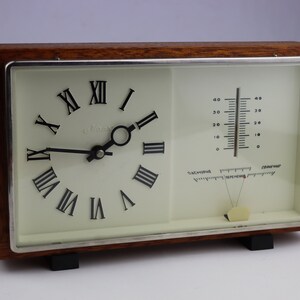 May include: A wooden framed clock with a white face and Roman numeral hour markers. The clock also has a thermometer and barometer.