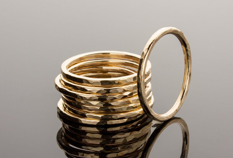 May include: A stack of ten gold hammered rings, with one ring laying beside the stack. The rings are all the same size and have a hammered texture.