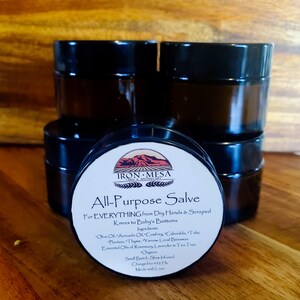 May include: A stack of dark brown glass jars with black lids. In front, a jar with a white label that reads "Iron Mesa All-Purpose Salve" with ingredients listed. The salve is for dry hands, scraped knees, and baby's bottoms.