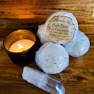 May include: A still-life image featuring a lit candle in a dark brown holder, several white bath bombs wrapped in clear plastic, and a clear quartz crystal. The bath bombs have a label that reads "Homebody Bath Bomb". The items are arranged on a wooden surface.