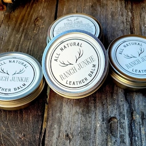 May include: Three silver tins of leather balm with a white label that reads "All Natural Ranch Junkie Leather Balm". The label features a stylized antler design.