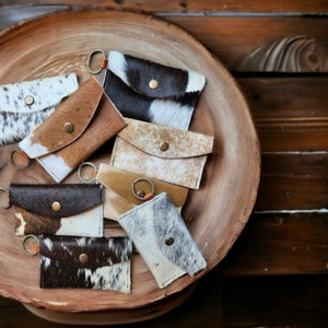 May include: A collection of small, leather keychains with a cowhide pattern in various colors, including brown, white, and black. Each keychain has a metal ring and a snap closure. The keychains are arranged on a wooden tray.