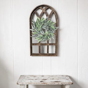 May include: A rustic, wooden wall decor piece featuring a faux green leaf wreath centered within a brown, arched window frame. The frame has a lattice design at the top and a grid pattern below. The decor is mounted on a white wall.