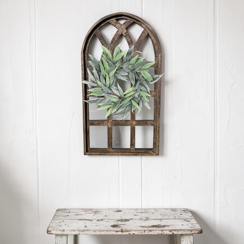 May include: A rustic, wooden wall decor piece featuring a faux green leaf wreath centered within a brown, arched window frame. The frame has a lattice design at the top and a grid pattern below. The decor is mounted on a white wall.