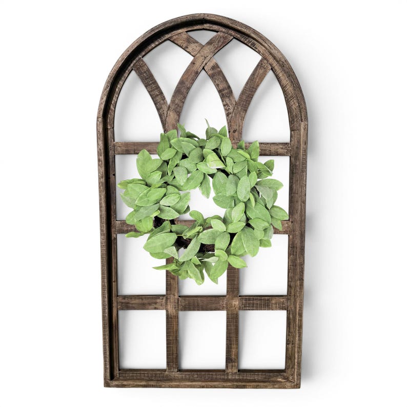 May include: A rustic, wooden window frame with an arched top and a lattice design. A circular wreath of green leaves is centered within the frame. The frame is a dark brown color, and the leaves are a soft green.