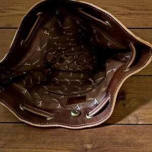 Puede incluir: Vista interior de un bolso de cuero marr&oacute;n con cord&oacute;n. El interior est&aacute; forrado con una tela estampada con un dise&ntilde;o de cornamenta repetido. El bolso tiene un cierre magn&eacute;tico y ojales met&aacute;licos para el cord&oacute;n.