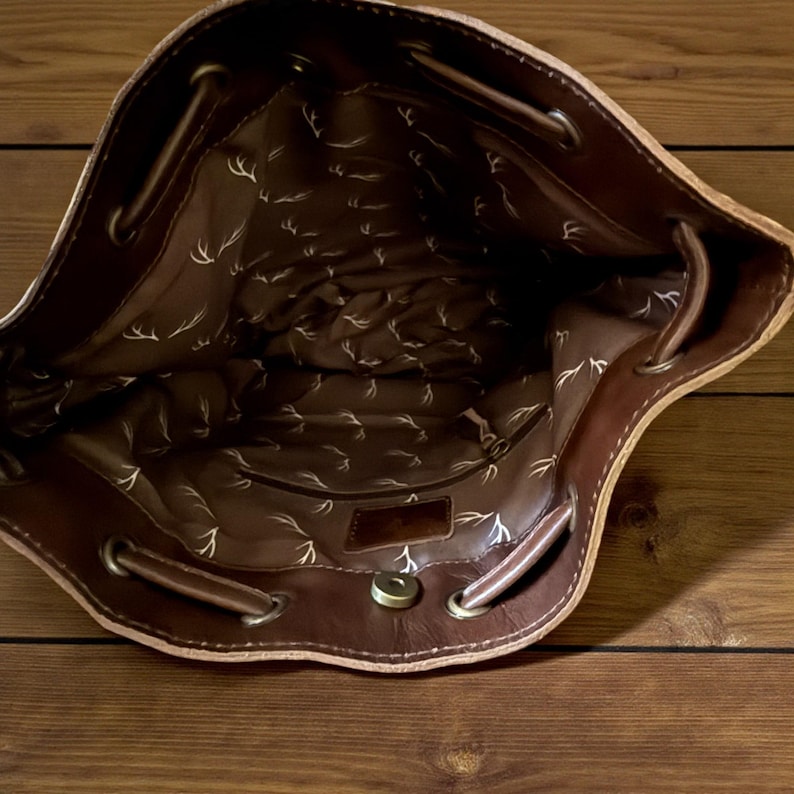 Puede incluir: Vista interior de un bolso de cuero marr&oacute;n con cord&oacute;n. El interior est&aacute; forrado con una tela estampada con un dise&ntilde;o de cornamenta repetido. El bolso tiene un cierre magn&eacute;tico y ojales met&aacute;licos para el cord&oacute;n.