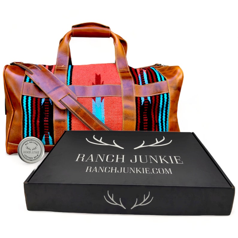 Op de afbeelding: Een bruine leren reistas met een kleurrijk geweven patroon. De tas heeft een lange band en staat op een zwarte doos met de tekst "RANCH JUNKIE" en "RANCHJUNKIE.COM".