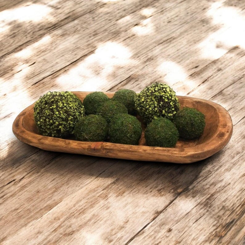 May include: A wooden bowl filled with green moss balls. The bowl is oval shaped and has a rustic finish.