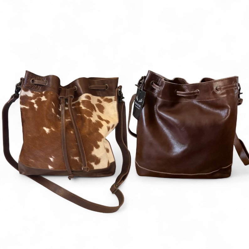 Puede incluir: Dos bolsos tipo cubo de cuero. Un bolso es marr&oacute;n con un estampado de piel de vaca en tonos marrones y crema. El otro bolso es de color marr&oacute;n liso. Ambos bolsos tienen cierres de cord&oacute;n, correas ajustables para el hombro y una etiqueta.