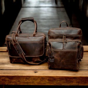 May include: Two brown leather bags, one a briefcase with a shoulder strap and the other a backpack with a top handle. Both bags have a distressed look and are made by the brand "Rider & Oak".