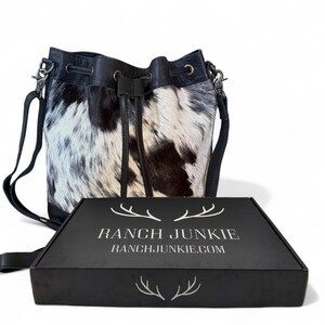 Puede incluir: Un bolso tipo cubo de piel de vaca en blanco y negro con cierre de cord&oacute;n y correa de cuero negro. El bolso est&aacute; junto a una caja negra con el texto "RANCH JUNKIE" y "RANCHJUNKIE.COM" en blanco.