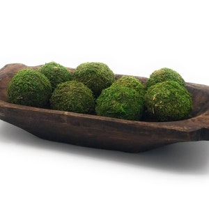 May include: A rustic wooden dough bowl filled with green moss balls. The bowl is dark brown and has a natural, uneven shape. The moss balls are a vibrant green and add a touch of nature to the bowl.