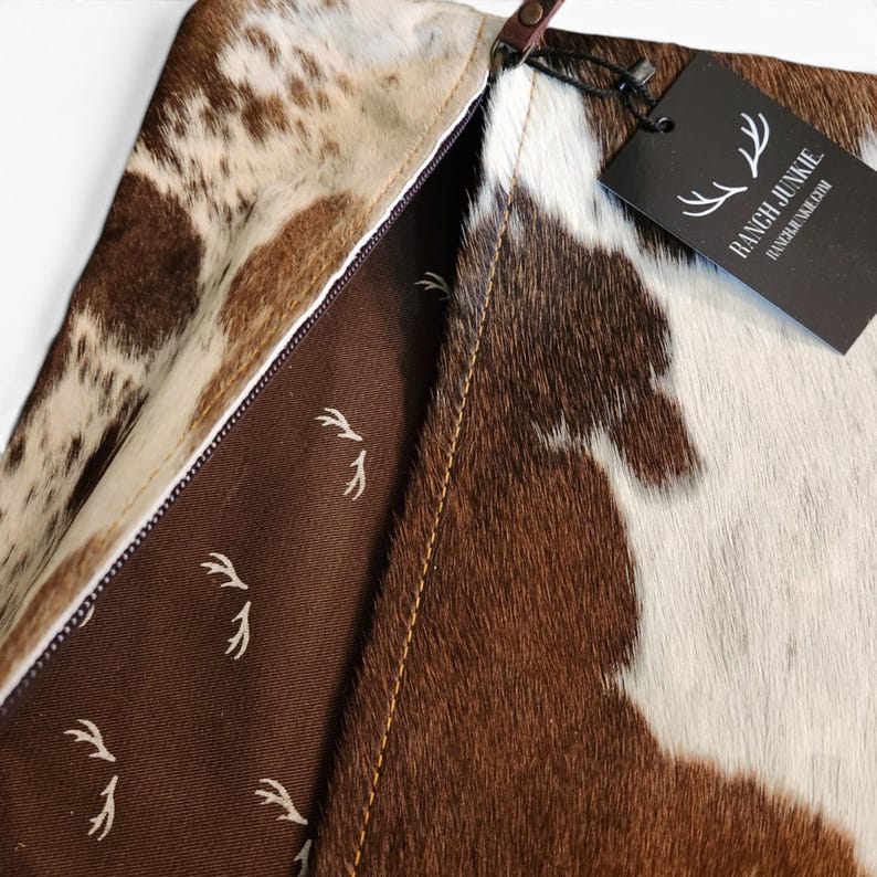 May include: A brown and white cowhide clutch with a zipper closure. The interior is lined with brown fabric featuring a white antler pattern. A black rectangular tag attached to the zipper pull displays the text RANCH JUNKIE and RANCHJUNKIE.COM.