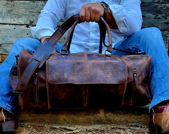 May include: A distressed brown leather duffel bag with a front pocket and shoulder strap. The bag has a worn, vintage look, with visible grain and texture. The bag is designed for travel and has a handle for carrying.