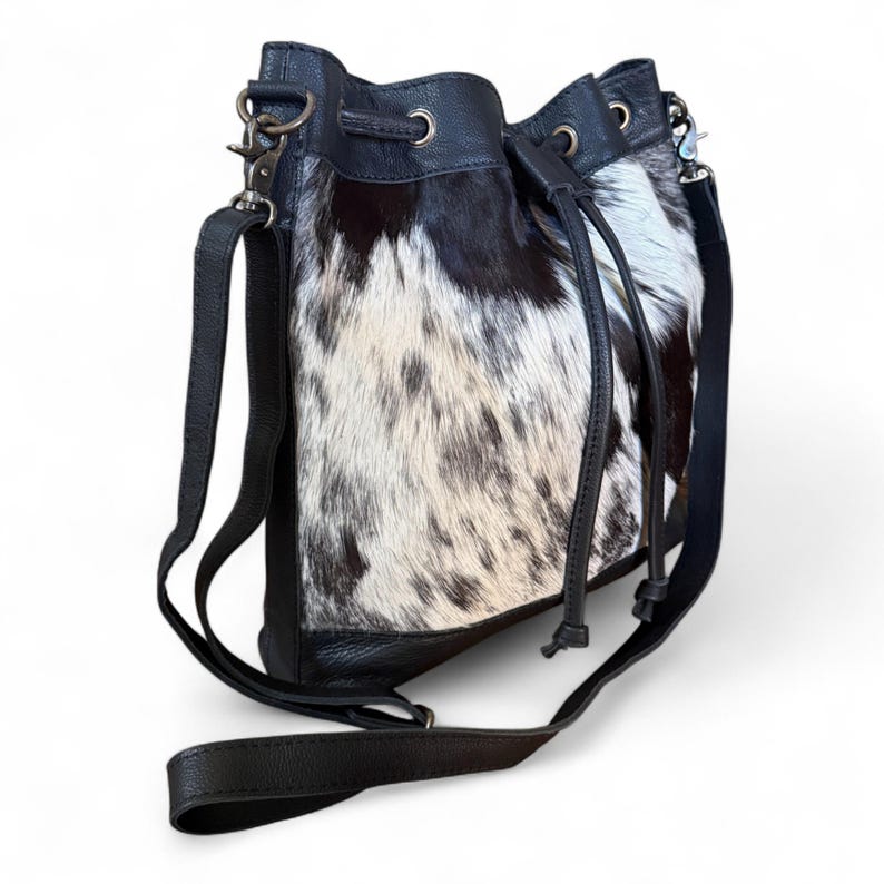Puede incluir: Bolso tipo cubo de piel de vaca en blanco y negro con cierre de cord&oacute;n de cuero negro y bandolera. El bolso tiene un cierre y ojales de metal plateado. El bolso est&aacute; dise&ntilde;ado para llevarse al hombro o cruzado.