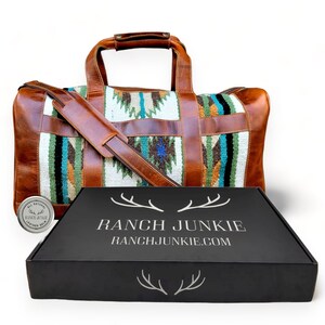 May include: A brown leather duffel bag with a colorful woven pattern and a leather strap. The bag is sitting on top of a black box with the text "RANCH JUNKIE" and "RANCHJUNKIE.COM" printed on it.