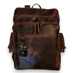 May include: A brown leather backpack with multiple pockets and zippers. The backpack has a tag that says "Handcrafted by Artisans" and "DrumSpeake".