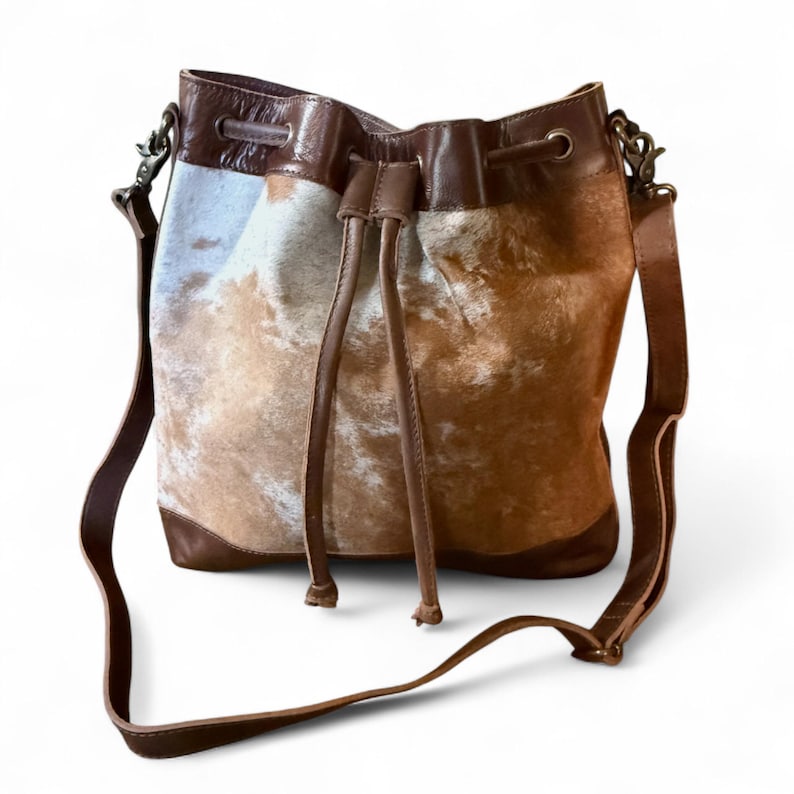 Puede incluir: Bolso tipo cubo con cierre de cord&oacute;n y bandolera de cuero marr&oacute;n. El bolso presenta un estampado de piel de vaca en tonos marr&oacute;n, beige y blanco. La base y los ribetes son de cuero marr&oacute;n oscuro, con herrajes color bronce.