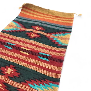 May include: A woven textile with a Southwestern design. The rug features a pattern of red, green, and turquoise geometric shapes and stripes. The top edge has a tan border with orange and gold stripes. The textile has a tasseled edge.