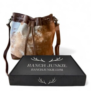 Puede incluir: Bolso tipo cubo de piel de vaca marr&oacute;n y blanca, con cierre de cord&oacute;n y correa de cuero. Junto a &eacute;l, una caja negra con el texto blanco "RANCH JUNKIE" y "RANCHJUNKIE.COM".