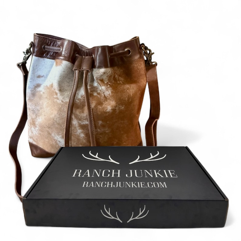 Puede incluir: Bolso tipo cubo de piel de vaca marr&oacute;n y blanca, con cierre de cord&oacute;n y correa de cuero. Junto a &eacute;l, una caja negra con el texto blanco "RANCH JUNKIE" y "RANCHJUNKIE.COM".