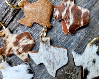 May include: Five leather keychains shaped like the state of Texas. Each keychain is made from a different cowhide pattern, including brown, white, and black. The keychains have gold metal key rings.