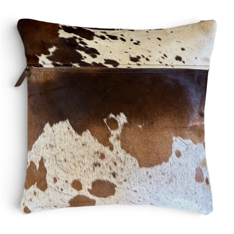 May include: A square throw pillow featuring a cowhide cover with a natural brown and white spotted pattern. The pillow is divided into two distinct sections by a horizontal seam, with a visible zipper pull located on the upper left side.