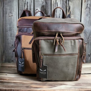May include: Two brown leather and canvas backpacks with a brown leather flap and a zipper closure. The backpacks have a brown leather tag with the text "RANCH JUNKIE" and a white deer antler logo.