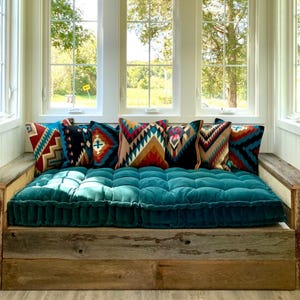 May include: A teal velvet tufted cushion sits on a wooden platform bed with a rustic wood frame. The bed is placed in front of a window with a view of trees. The bed is decorated with colorful patterned throw pillows.