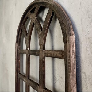 May include: A rustic, arched wooden window frame with a grid pattern. The frame is made of weathered, dark brown wood, showcasing visible grain and texture. The design features a series of intersecting lines, creating a geometric pattern. The overall aesthetic is vintage and farmhouse-inspired.