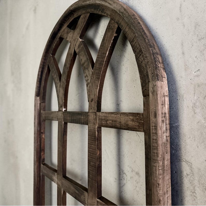 May include: A rustic, arched wooden window frame with a grid pattern. The frame is made of weathered, dark brown wood, showcasing visible grain and texture. The design features a series of intersecting lines, creating a geometric pattern. The overall aesthetic is vintage and farmhouse-inspired.
