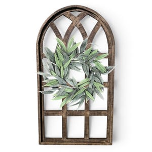 May include: A decorative wooden window frame with a green and gray leaf wreath. The frame is arched at the top and has a dark brown finish. The wreath is centered on the frame and is made of faux leaves.