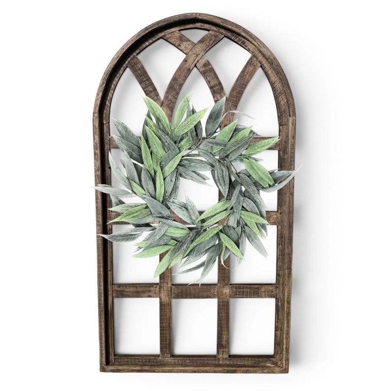 May include: A decorative wooden window frame with a green and gray leaf wreath. The frame is arched at the top and has a dark brown finish. The wreath is centered on the frame and is made of faux leaves.
