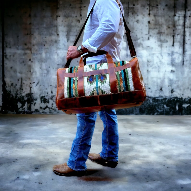 May include: A brown leather duffel bag with a colorful woven pattern. The bag has a long strap and is being carried by a person wearing blue jeans.
