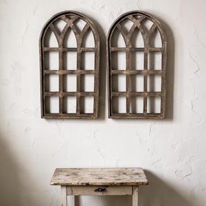 May include: Two arched wooden window frames with a grid pattern are mounted on a white wall. The frames have a distressed, rustic finish. Below, a small, weathered wooden table with a single drawer is visible.