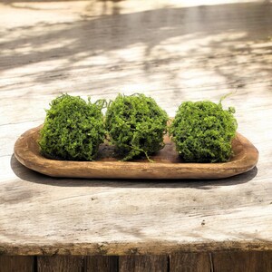 May include: Three green moss balls arranged in a wooden oval tray. The tray is on a wooden surface.