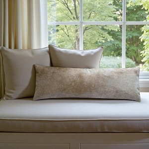 May include: Three decorative pillows in neutral tones rest on a window seat. The pillows include a large square pillow, a smaller rectangular pillow, and a long, lumbar pillow. The lumbar pillow has a textured, mottled appearance. The window behind the pillows looks out onto a green, leafy landscape.