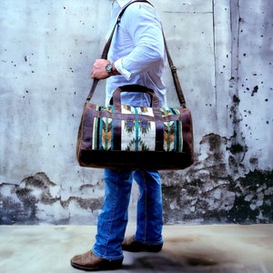 May include: A brown leather duffel bag with a colorful woven pattern. The bag has a long strap and is being carried by a person wearing blue jeans.