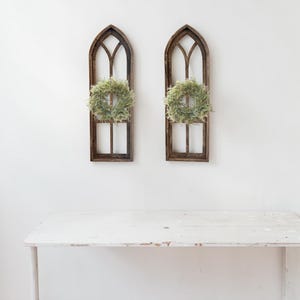 May include: Two wooden arched window frames with small green wreaths hanging inside. The frames are a dark brown color and are mounted on a white wall. A white table is in the foreground.