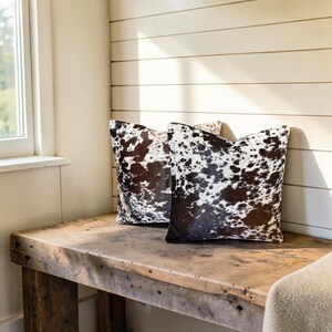 May include: Two square throw pillows featuring a natural cowhide pattern with irregular patches of dark brown and white fur. The pillows are positioned side by side, displaying a textured, organic hide appearance with varied spot sizes and shapes.