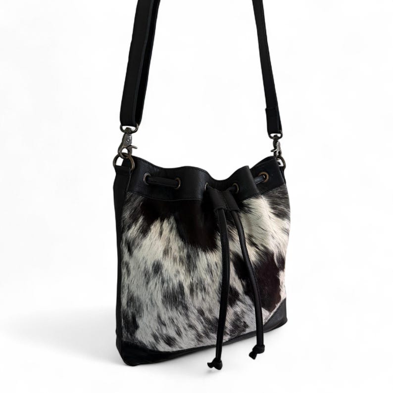 Puede incluir: Un bolso tipo cubo de piel de vaca en blanco y negro con cierre de cord&oacute;n. El bolso tiene una base, ribete y correa de hombro ajustables de cuero negro. El bolso est&aacute; suspendido sobre un fondo blanco.