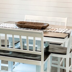 May include: White farmhouse style dining table with a wooden top and white chairs. A wooden bowl sits on the table with a white and brown runner.