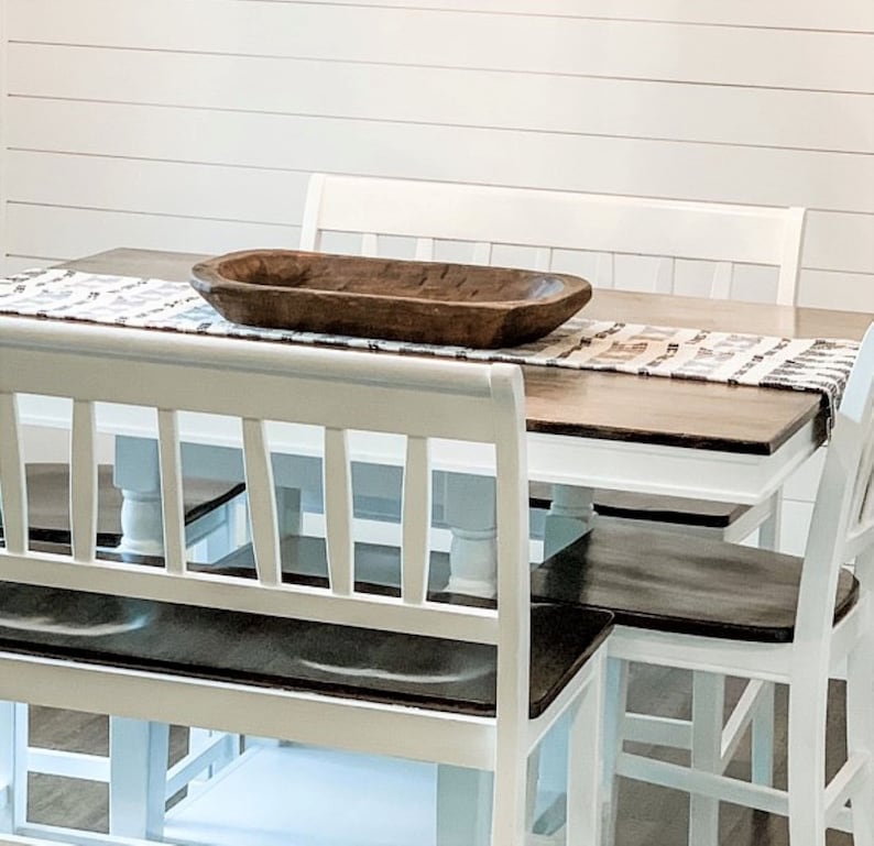 May include: White farmhouse style dining table with a wooden top and white chairs. A wooden bowl sits on the table with a white and brown runner.