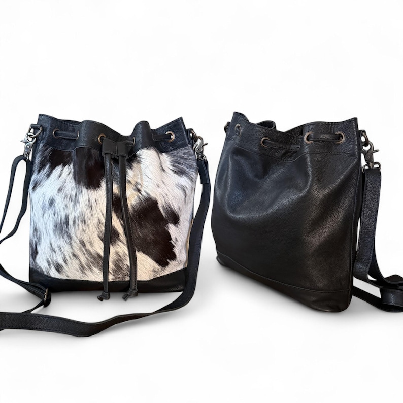 Puede incluir: Dos bolsos tipo cubo. Uno es de piel de vaca en blanco y negro con ribete de cuero negro y cierre de cord&oacute;n. El otro es de cuero negro liso con una correa a juego y cierre de cord&oacute;n. Ambos tienen herrajes plateados.