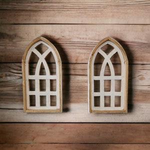 May include: Two white wooden arched window wall decor pieces with a gold trim. The windows are on a wood background.