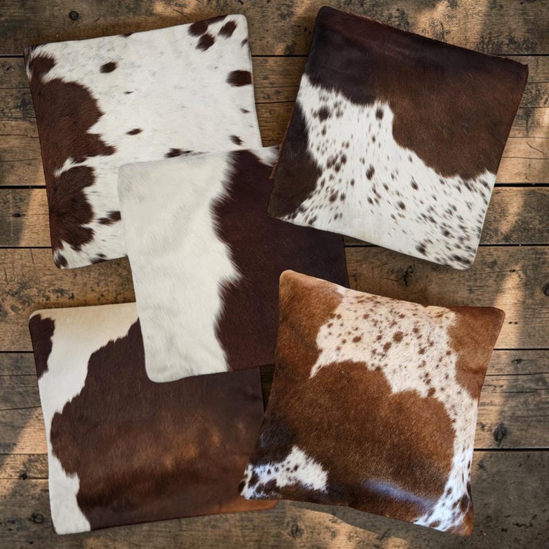 May include: A collection of four square throw pillows featuring natural cowhide patterns in various shades of brown, tan, and white. Each pillow displays a unique arrangement of patches, spots, and solid color blocks across the surface.