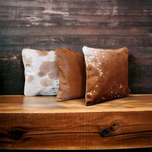 May include: Three square decorative pillows with cowhide covers in shades of brown and white. The pillows are arranged on a wooden surface against a dark wood background. The cowhide patterns vary, adding texture and visual interest to the home decor.